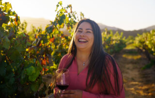 smiling woman pioneers of california's vineyard industry holding red wine outdoors amongst grapevines at sunset.